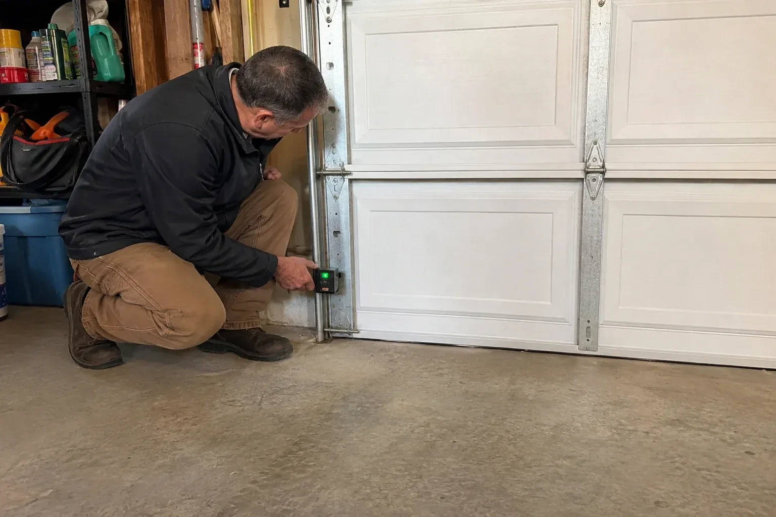 Technician kneeling beside a white paneled garage door, holding a small black box with a green light against the vertical track. — Photo-Eye Sensor Alignment or Replacement, Haldimand County